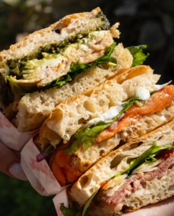 A stack of focaccia sandwiches showing fillings of pesto, burrata, salmon, beef and arugula